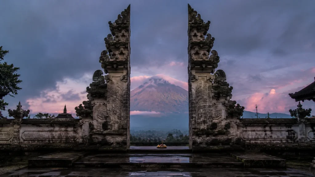 Ornate Balinese split temple gate at dawn with Mount Agung in the distance and frangipani offering at the base