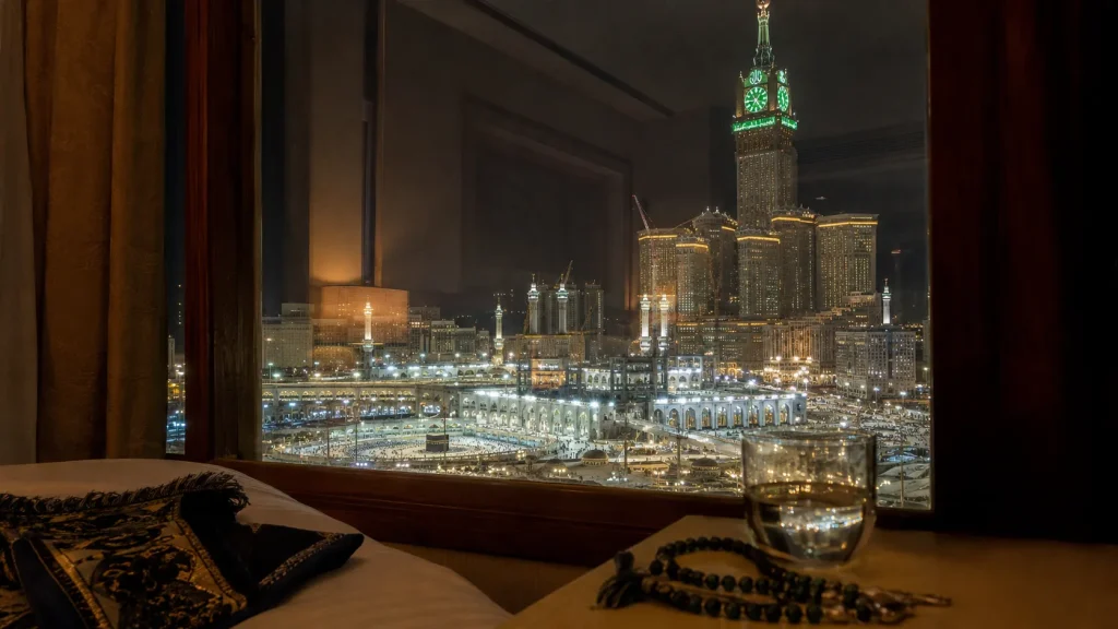 Hotel room in Makkah with prayer mat and tasbih on bed, window view of illuminated Masjid al-Haram at night | Umrah from Pakistan