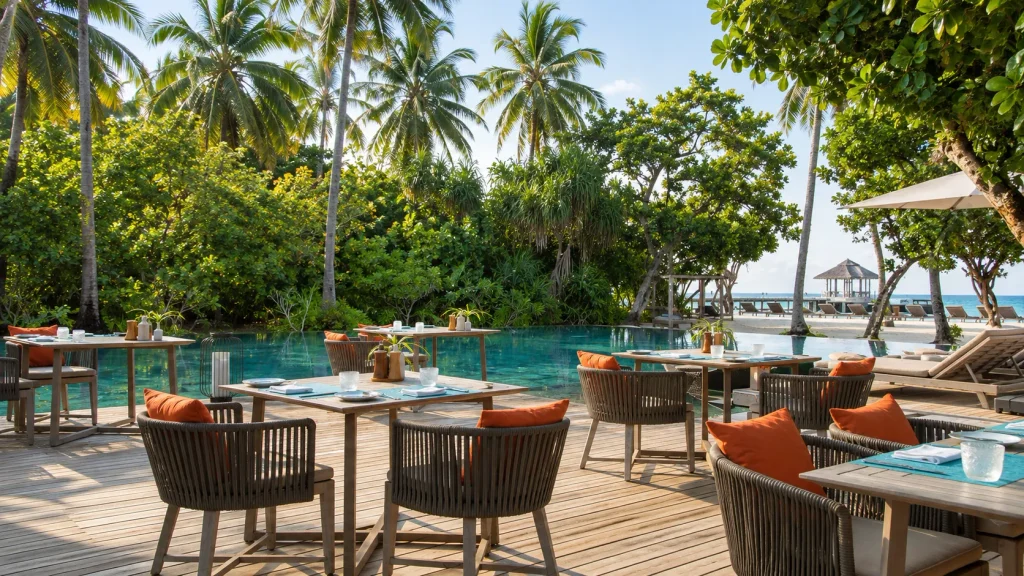 Palm-shaded outdoor dining deck beside a lagoon pool at a Maldives resort, with beach loungers visible beyond.
